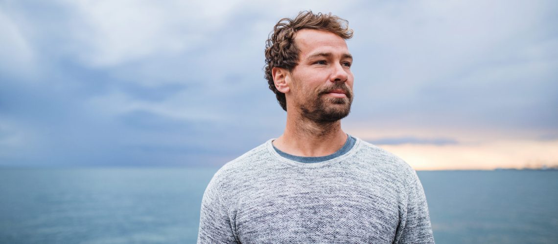 Portrait of mature man standing outdoors on beach at dusk.