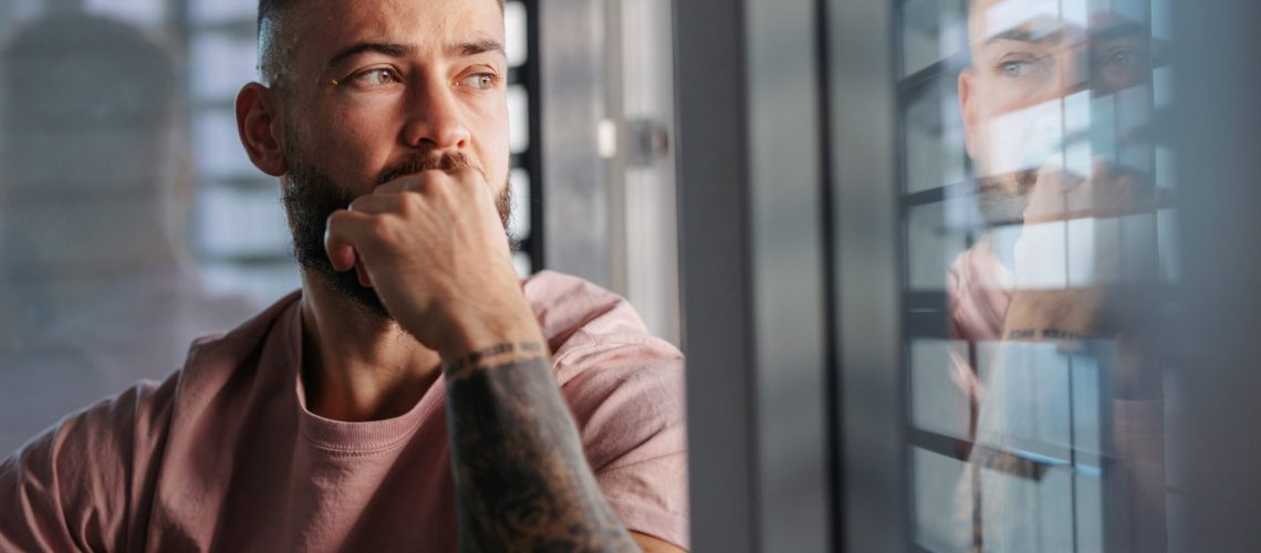 Handsome calm man in pink t-shirt with tattoo on arms looking through window.